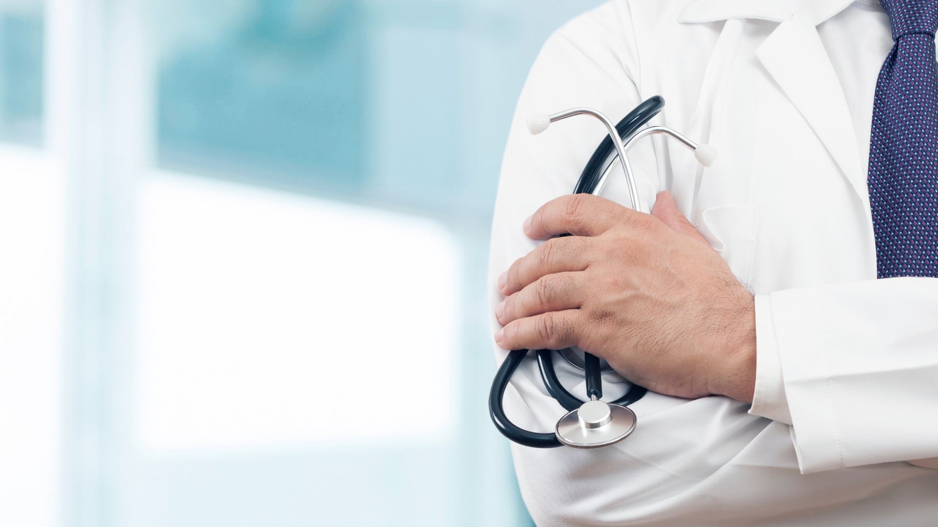 Plano aproximado de um médico com bata branca e gravata, de braços cruzados, a segurar um estetoscópio. O fundo é desfocado e claro.
